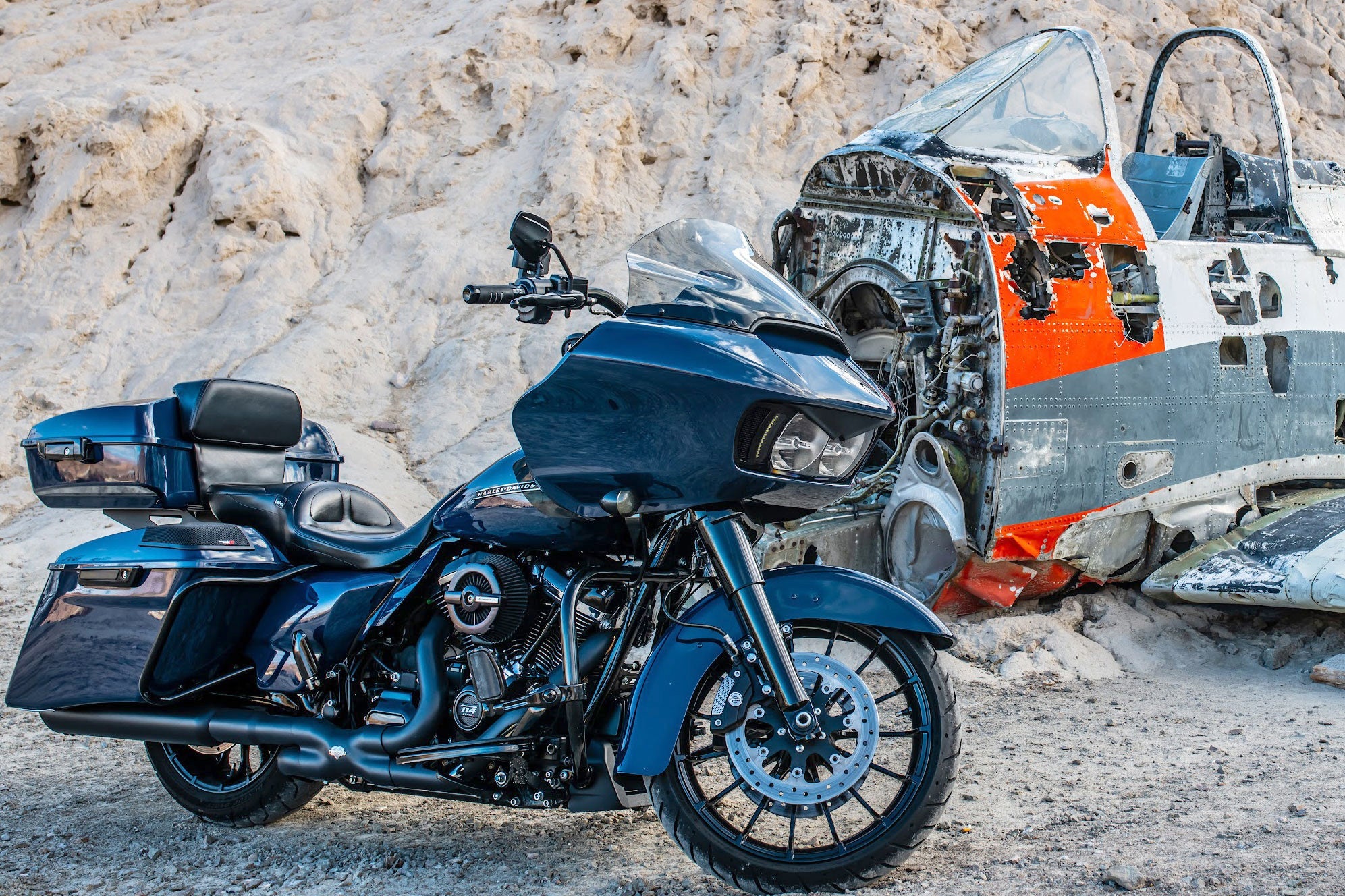 A 2014-2023 Harley Davidson® Cut In Lid Kit with ST692CX2 (2Ω) by Cerwin Vega stands on rocky ground, while behind it lies a worn airplane cockpit painted in white and orange. The sleek new design of the motorcycle kit contrasts strikingly with the aged cockpit.