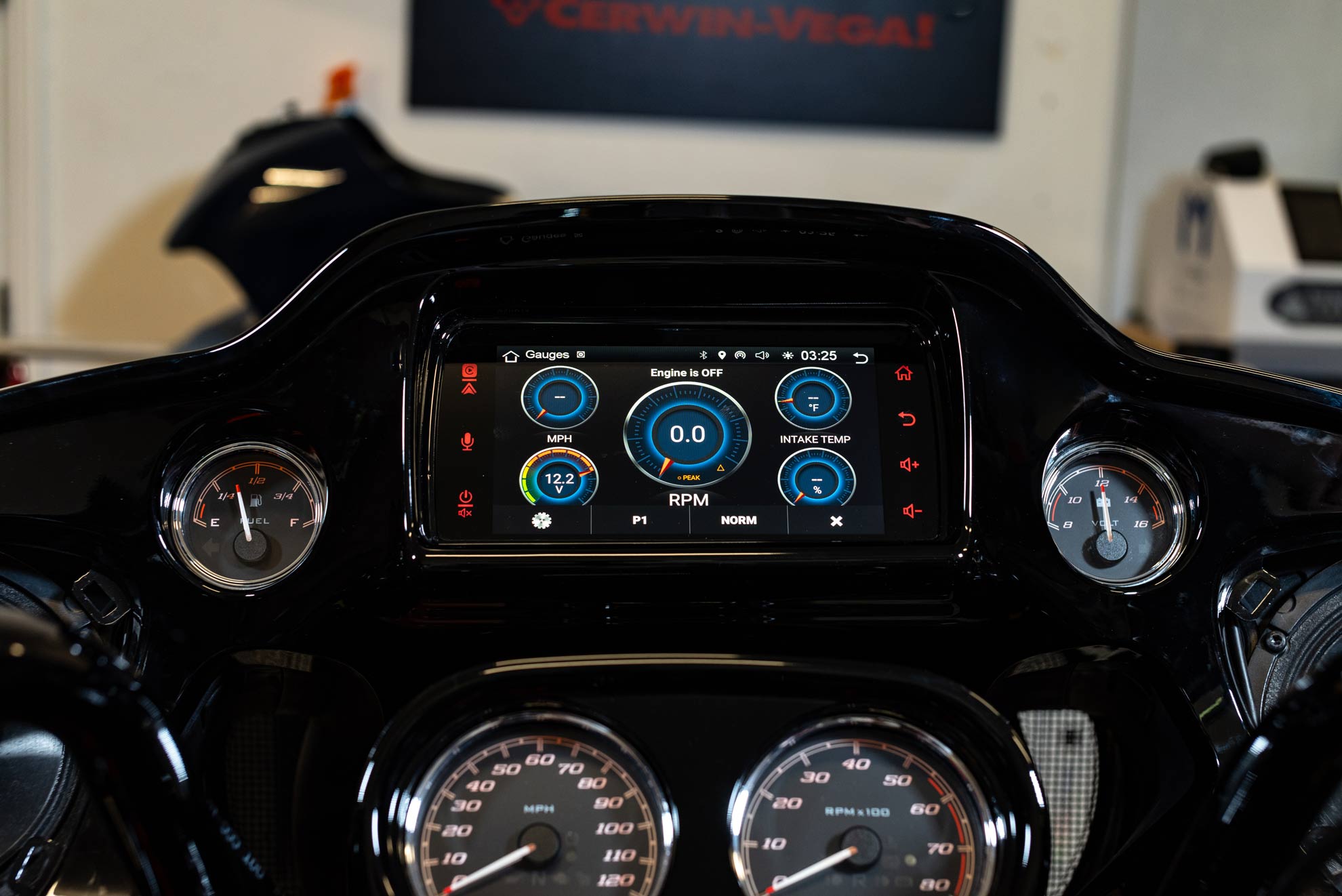 Close-up of a Harley Davidson motorcycle dashboard featuring the 2014-2023 Harley Davidson® Plug N-Play Media Receiver RPMHD14, showcasing digital RPM, mileage, and temperature gauges with illuminated blue and white graphics alongside analog speedometers. A blurred Cerwin-Vega sign is visible in the background.