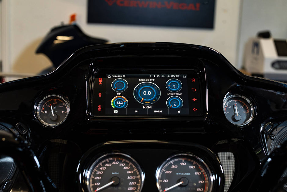 Close-up of a Harley Davidson motorcycle dashboard featuring the 2014-2023 Harley Davidson® Plug N-Play Media Receiver RPMHD14, showcasing digital RPM, mileage, and temperature gauges with illuminated blue and white graphics alongside analog speedometers. A blurred Cerwin-Vega sign is visible in the background.