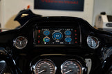 Close-up of a Harley Davidson motorcycle dashboard featuring the 2014-2023 Harley Davidson® Plug N-Play Media Receiver RPMHD14, showcasing digital RPM, mileage, and temperature gauges with illuminated blue and white graphics alongside analog speedometers. A blurred Cerwin-Vega sign is visible in the background.