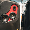 Close-up of a car door panel featuring Cerwin Vega's Stroker Pro CVMPCL6.5 audio system against a red background, showcasing three circular speakers for exceptional clarity, with the largest at the bottom. The open door reveals part of the car's interior and a blue sky outside.