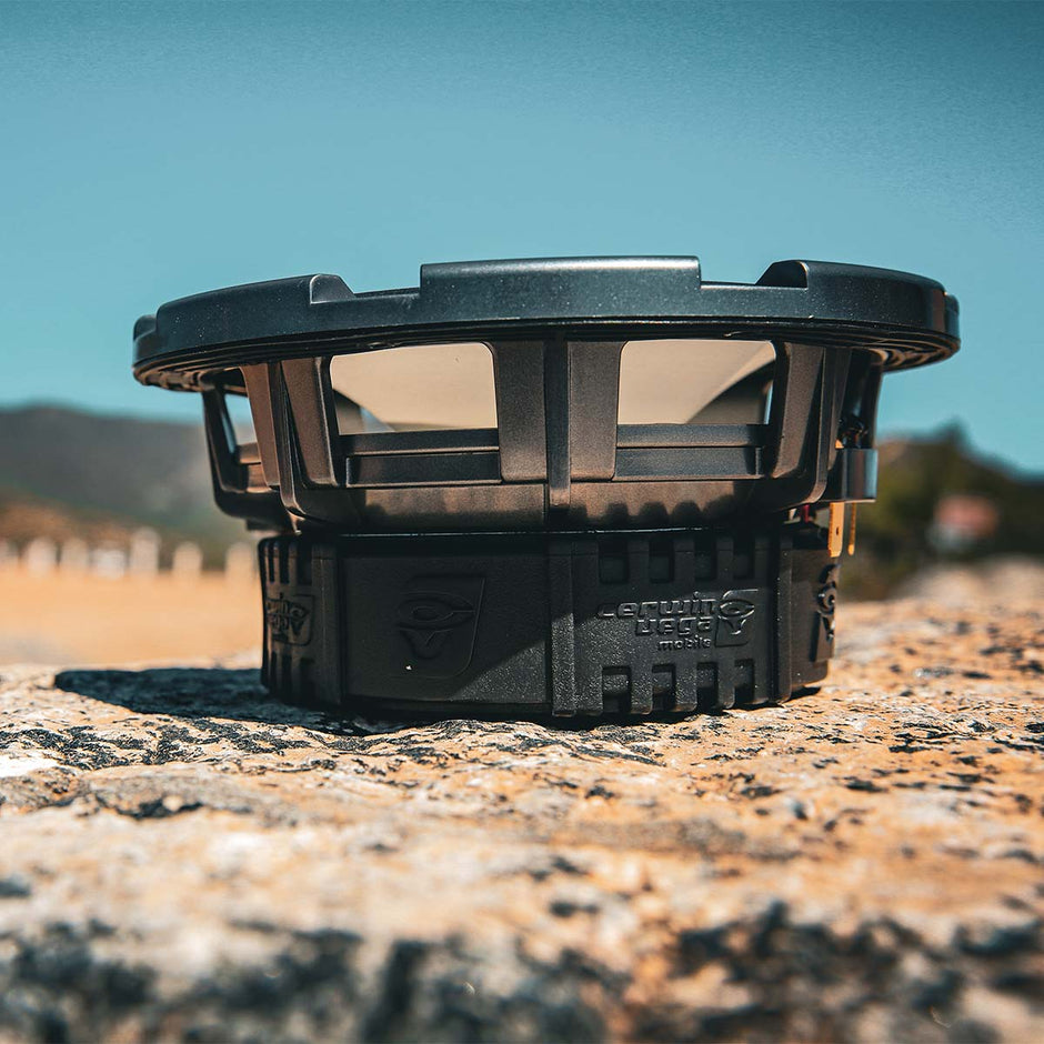Close-up of the RPM Stroker SM65F4 black car subwoofer with weather resistance on a textured rocky surface. The design highlights the brand logo and enclosure details, set against a clear blue sky with distant blurred hills, suggesting an outdoor setting.